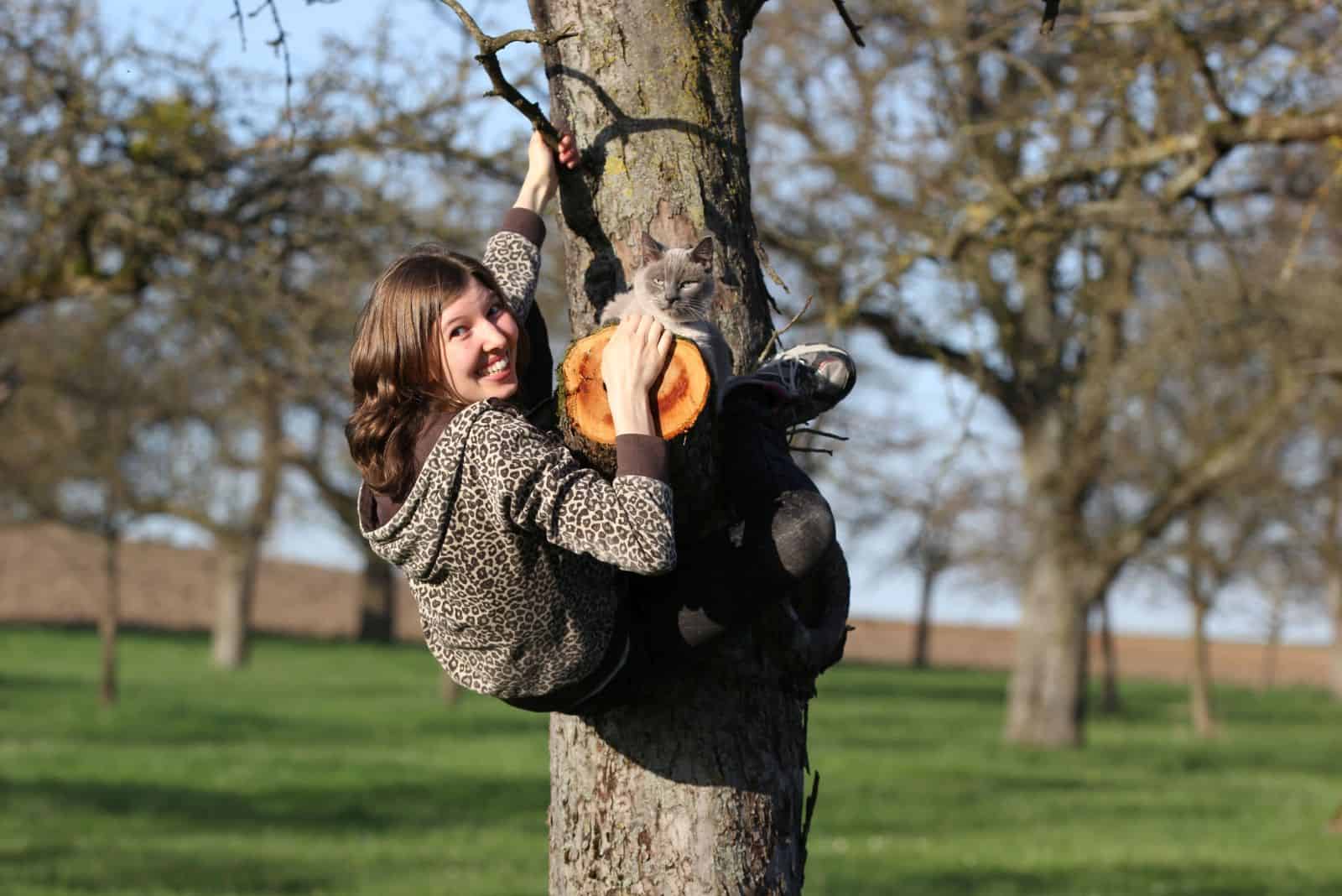 a girl climbs a tree while a gray cat sits on it