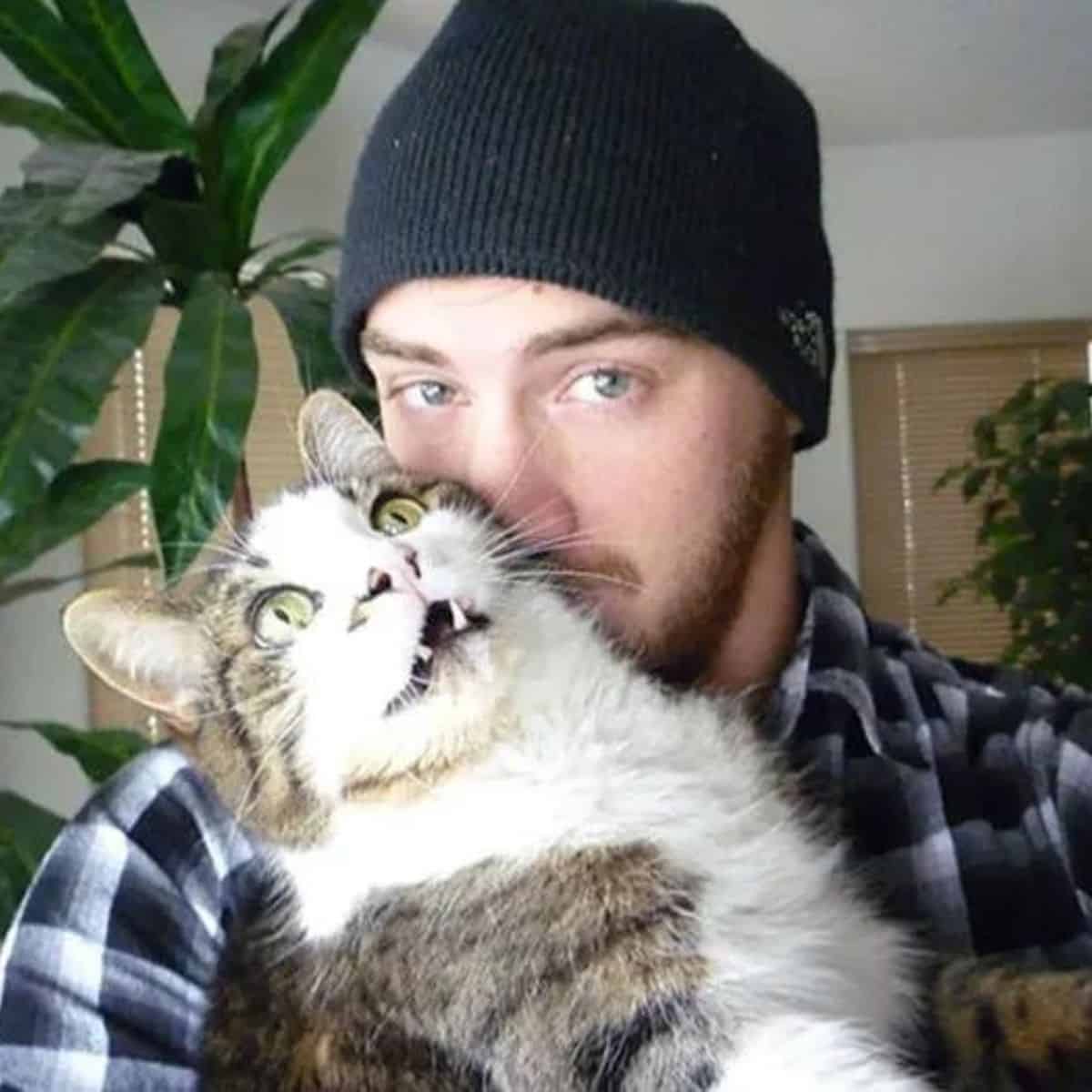 a man with a cap on his head holds a cat in his arms